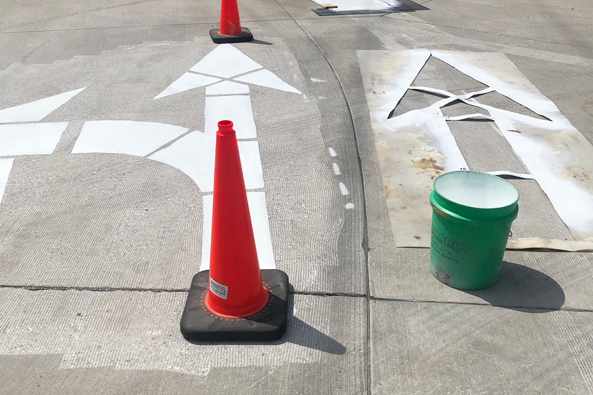 Orange cones protect freshly painted large arrows on a street. A stencil lays nearby.