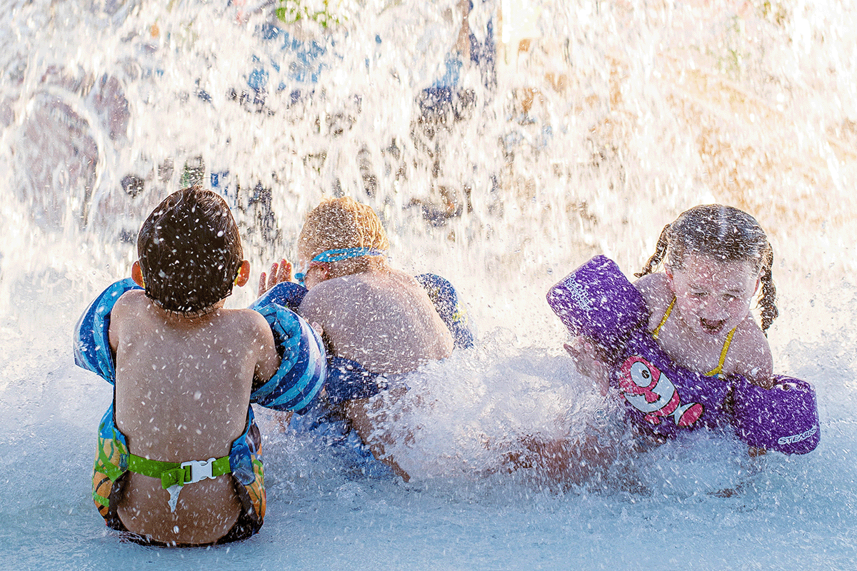 Kids in the outdoor pool, all wearing water wings, squeal with delight as they are splashed with water from above.