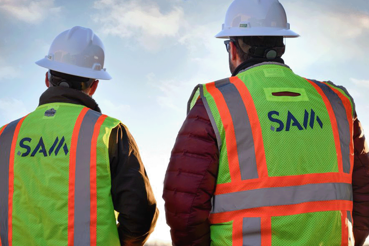 A pair of SAM employees in safety vests and hard hats against a blues sky.