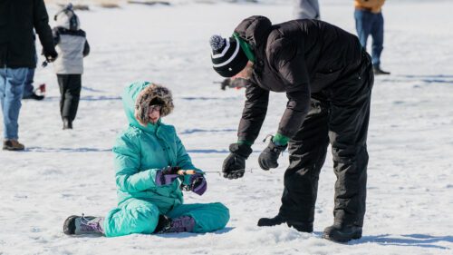 A well-bundled child prepares to ice fish with help of an adult while others walk around in the background.
