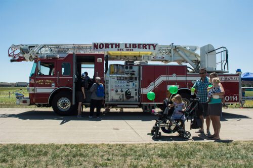 A North Liberty Fire Department ladder truck on display at Centennial Park as a family holding green balloons walks by.