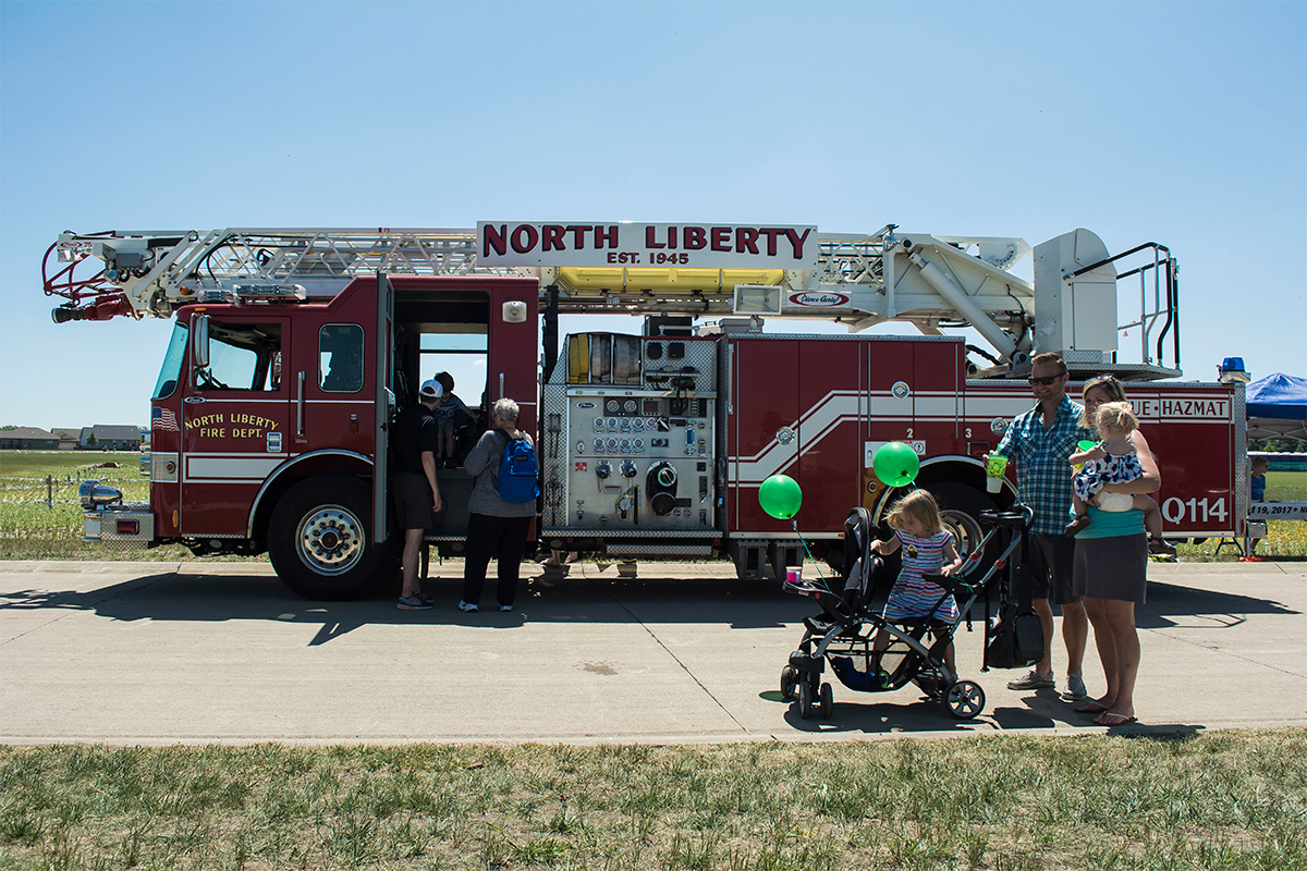 A North Liberty Fire Department ladder truck on display at Centennial Park as a family holding green balloons walks by.