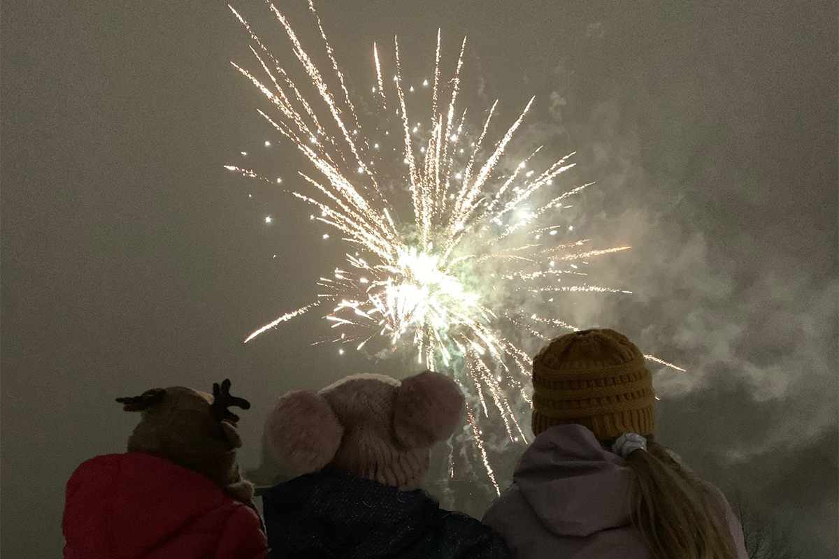 Three children bundled in snow gear watch fireworks burst overhead.