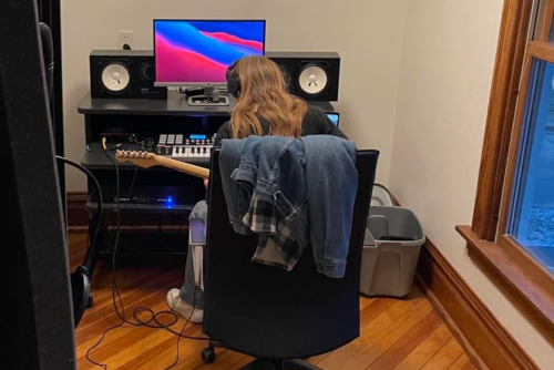 A teen with long hair, wearing headphones, sits in a chair with their back to the camera, in front of a computer with large speakers, a music keyboard and playing a guitar. Their jean jacket is laying over the back of the chair.