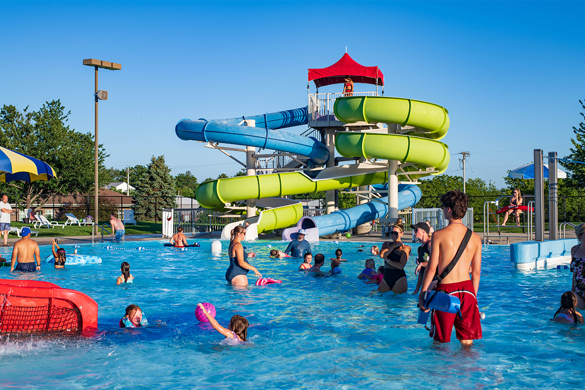 Kids swim in an outdoor pool while a lifeguard in red swimming trucks watches. Bright green and blue water slides are in the background.